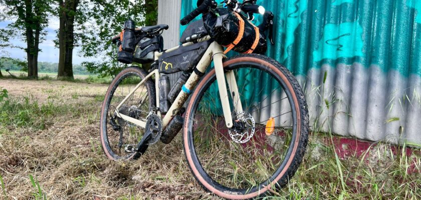 Scott Speedster Gravel bicycle, beige, with bikepacking bags attached to handlebar, frame and saddle. Bike is leaning on a abandoned industrial building wall with a graffiti in a field.